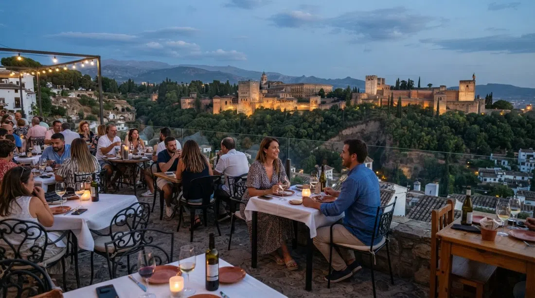 terraza panorámica de Casa Juanillo sobre Sacromonte, con la Alhambra llenando la vista tras el cristal y la luz del atardecer sobre las mesas