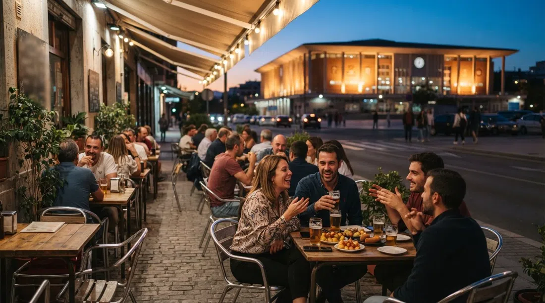 la terraza al atardecer de Cervecería Ecu con locales tomando cañas y tapas enormes, el Palacio de Deportes brillando al otro lado de la calle tras la puesta de sol